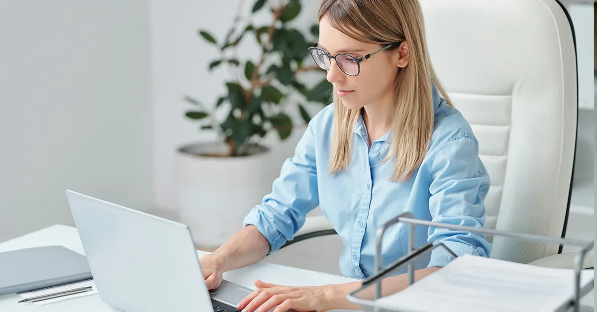 Individual sitting at a white desk in a modern office, typing on a laptop with documents and a file tray nearby.