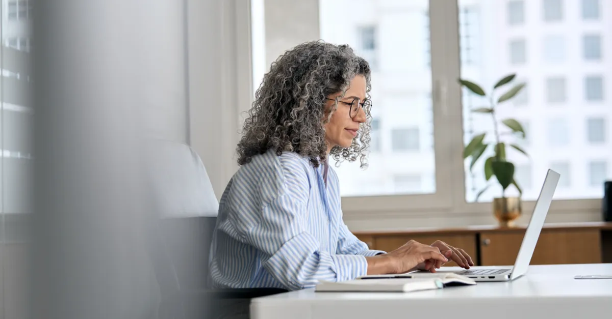 Person sitting at a desk, holding a pen and looking at a laptop while writing in a notebook.