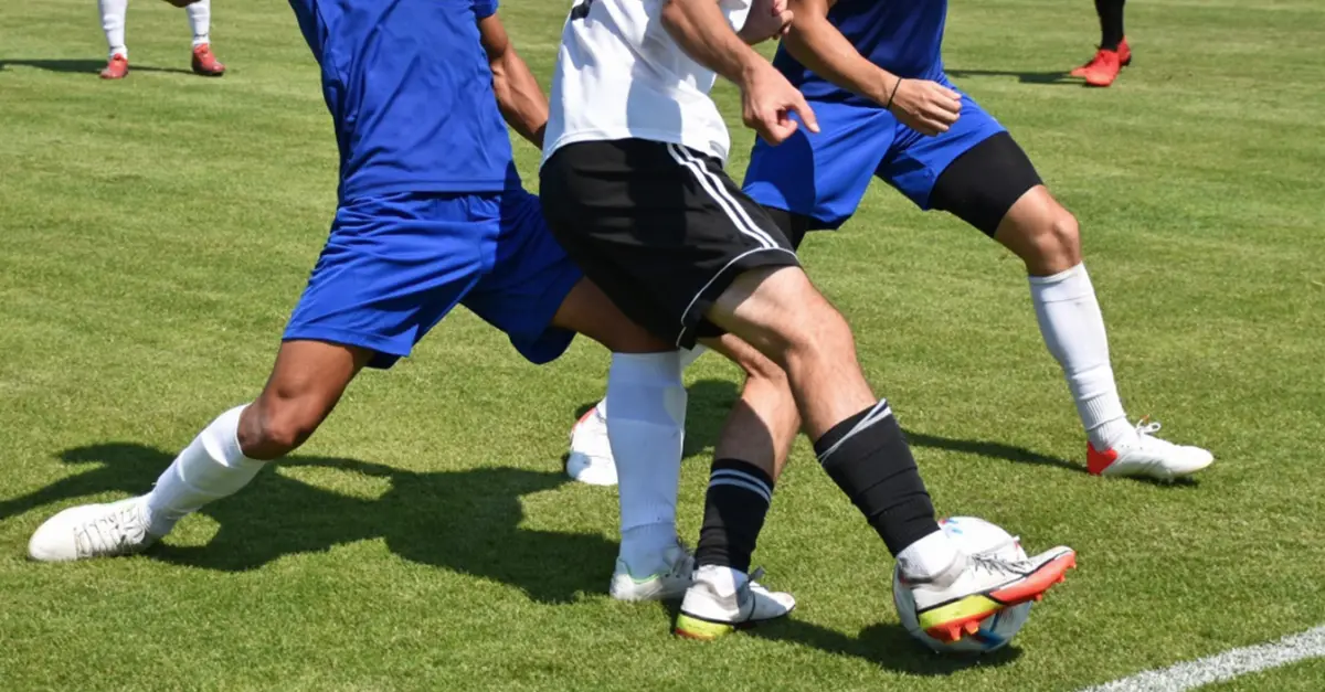 Multiple players on a grass field closely contesting possession of a soccer ball, with legs positioned around the ball during active play.