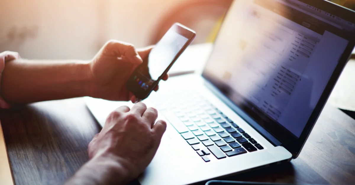 Hands holding a smartphone while using a laptop at a desk, with sunlight shining across the workspace.