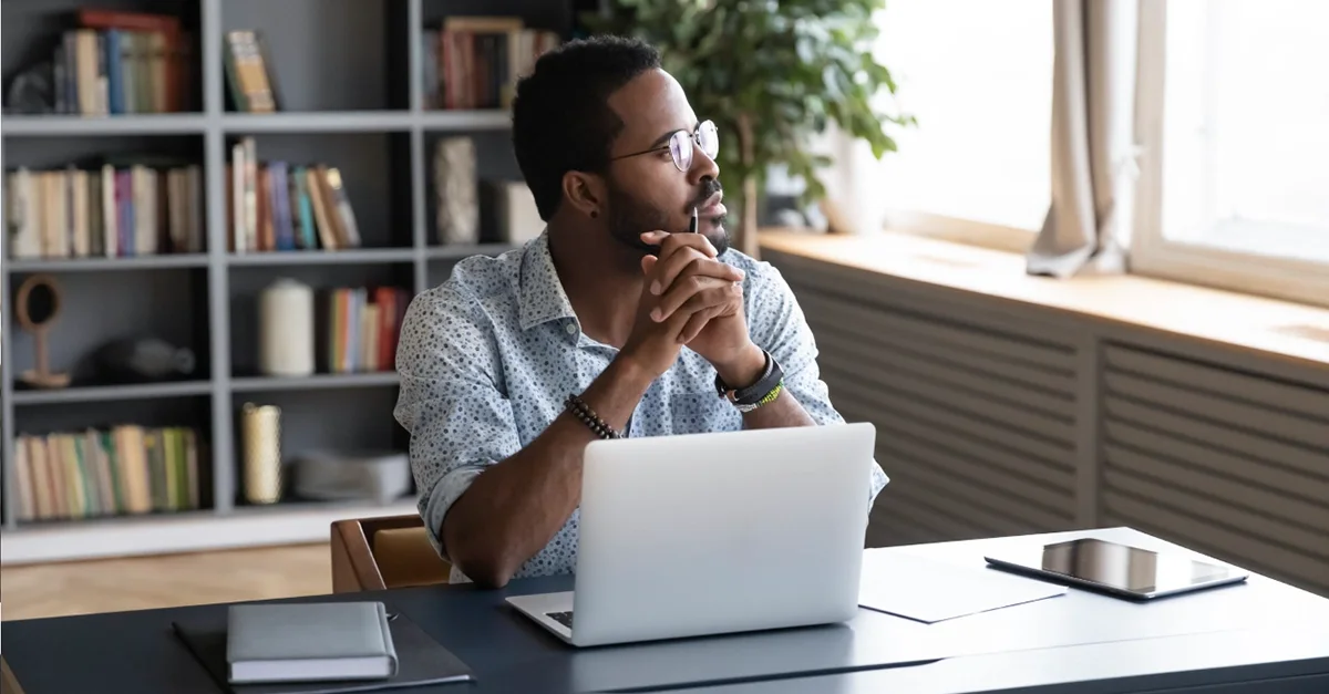 Individual seated at a desk in a home office or study area, resting hands together while looking away from a laptop, with notebooks and a tablet on the desk and bookshelves in the background.