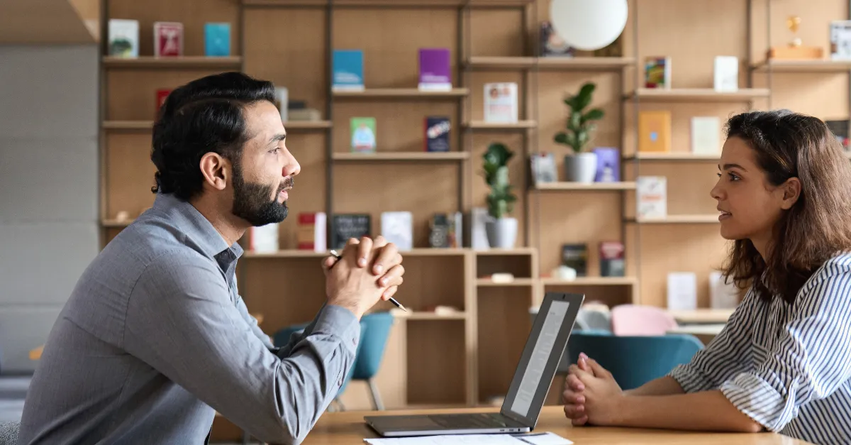 Two colleagues sitting across a table having a discussion in a modern office with shelves in the background.