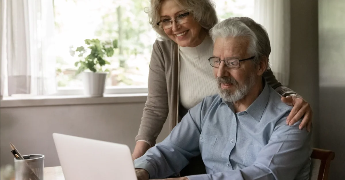 Two individuals sitting close together at a table, looking at a laptop screen in a bright room with large windows and indoor plants.