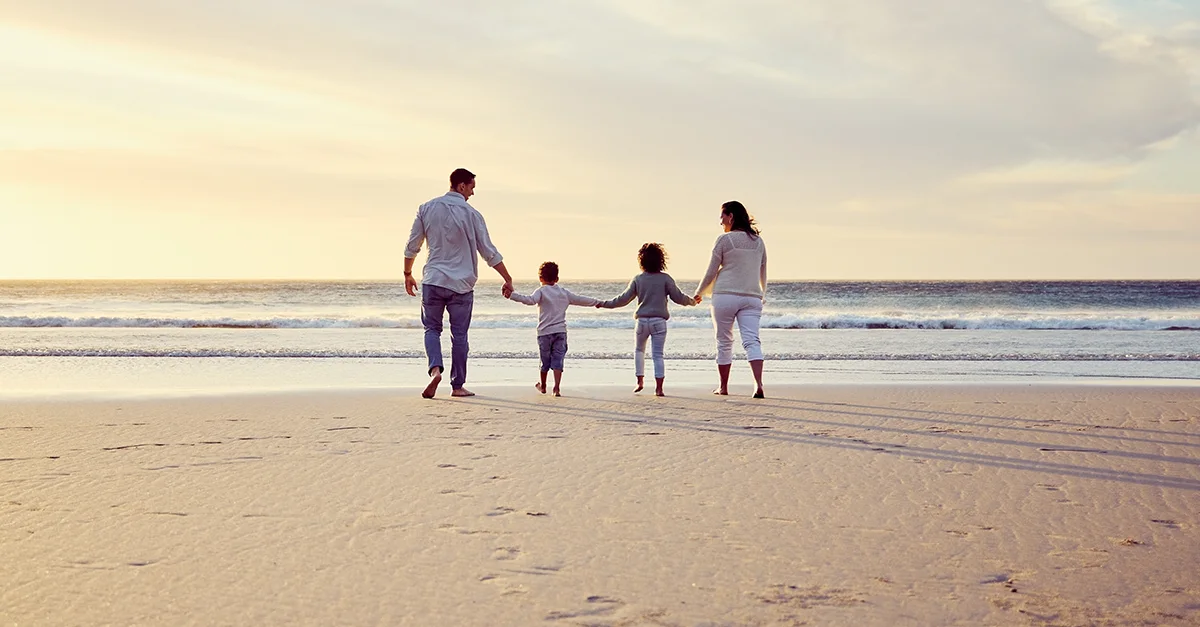 Four people walking hand in hand along a sandy beach at sunset, with gentle waves in the background.