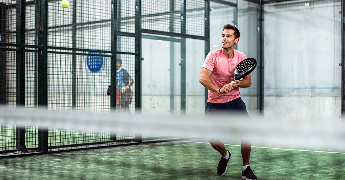 Individual on an indoor padel court preparing to return a shot with a padel racket, with a ball visible in the air near the net.