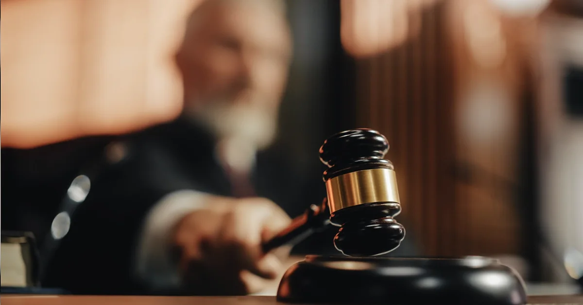 A judge’s hand holding a gavel and striking it on a sounding block in a courtroom setting.