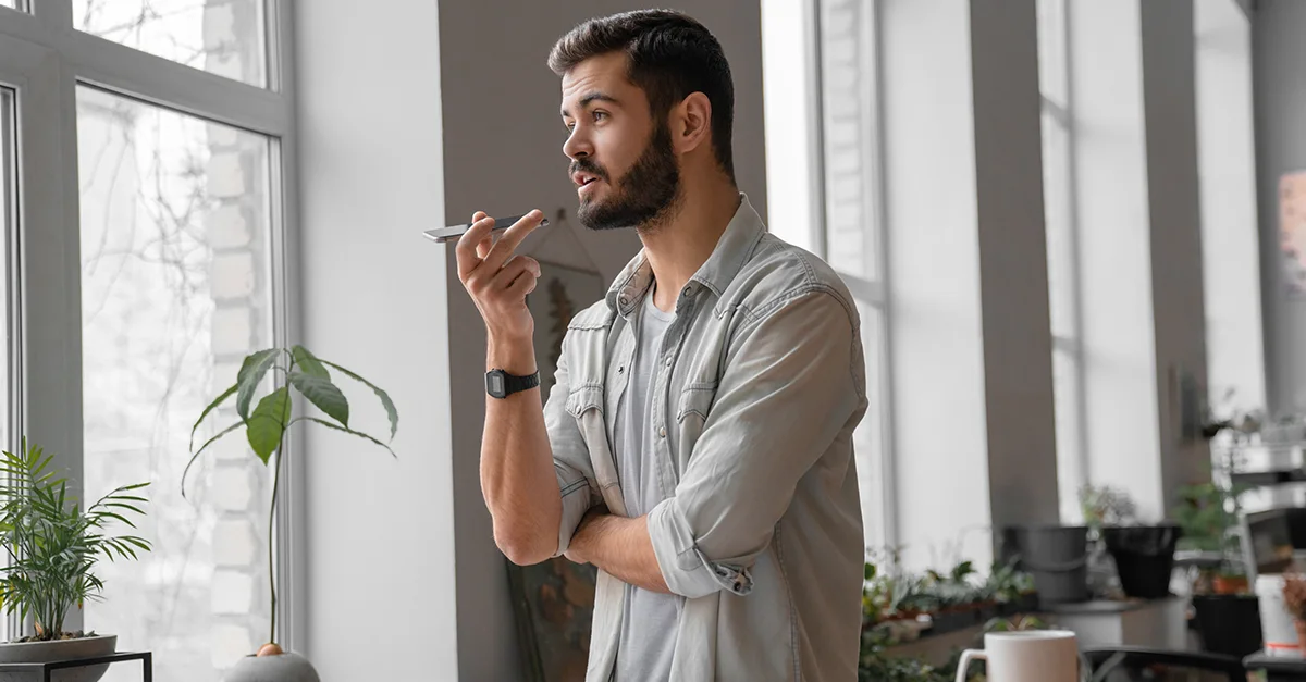 Individual standing indoors and holding a smartphone near the mouth as if recording or sending a voice message, with one arm crossed and windows in the background.