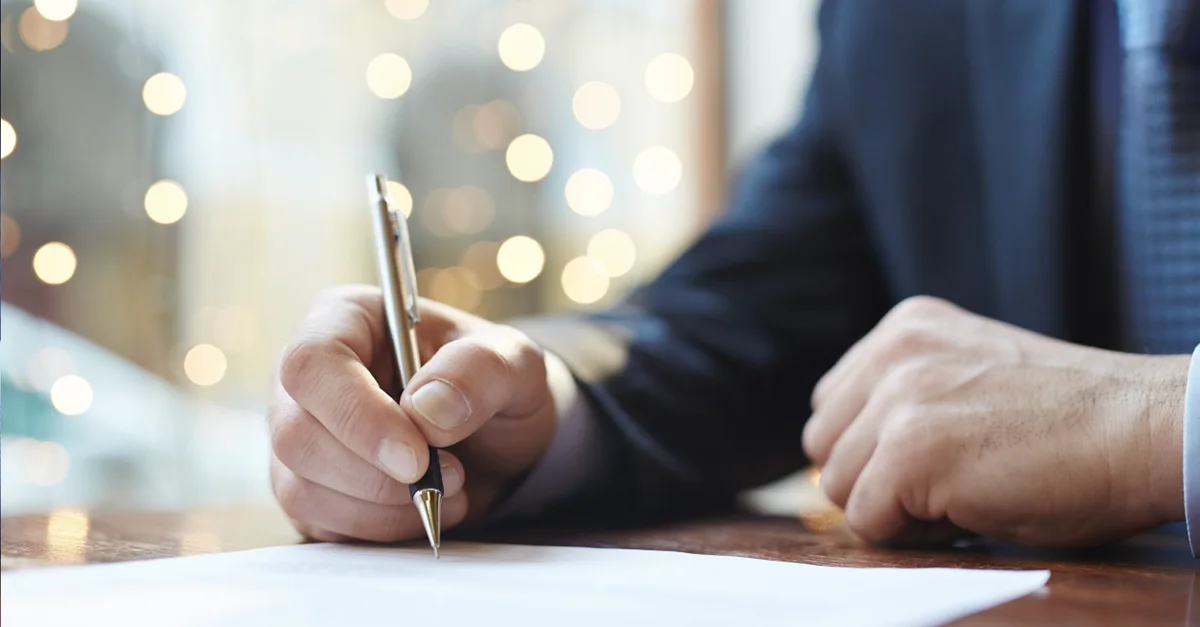Close‑up of a person in business attire signing a document with a pen at a desk.