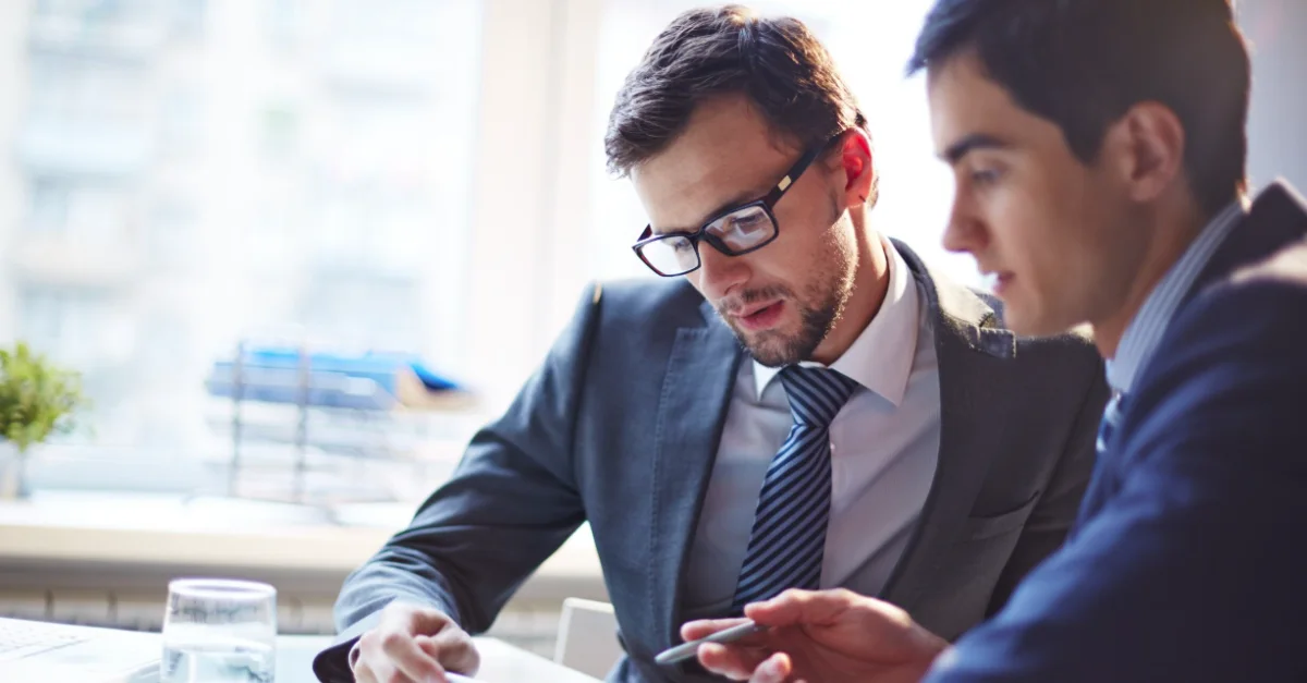 Two colleagues in a professional office setting sitting at a desk and reviewing information on a tablet during a discussion