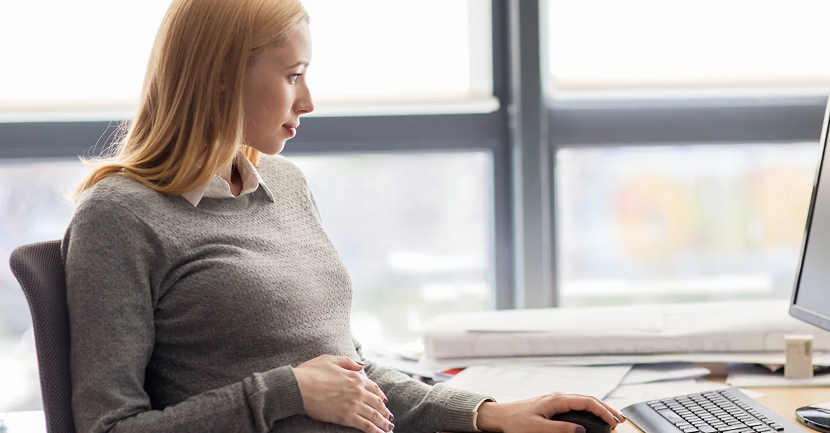 Individual seated at an office desk with one hand on the abdomen, working on a keyboard with documents and office supplies nearby.