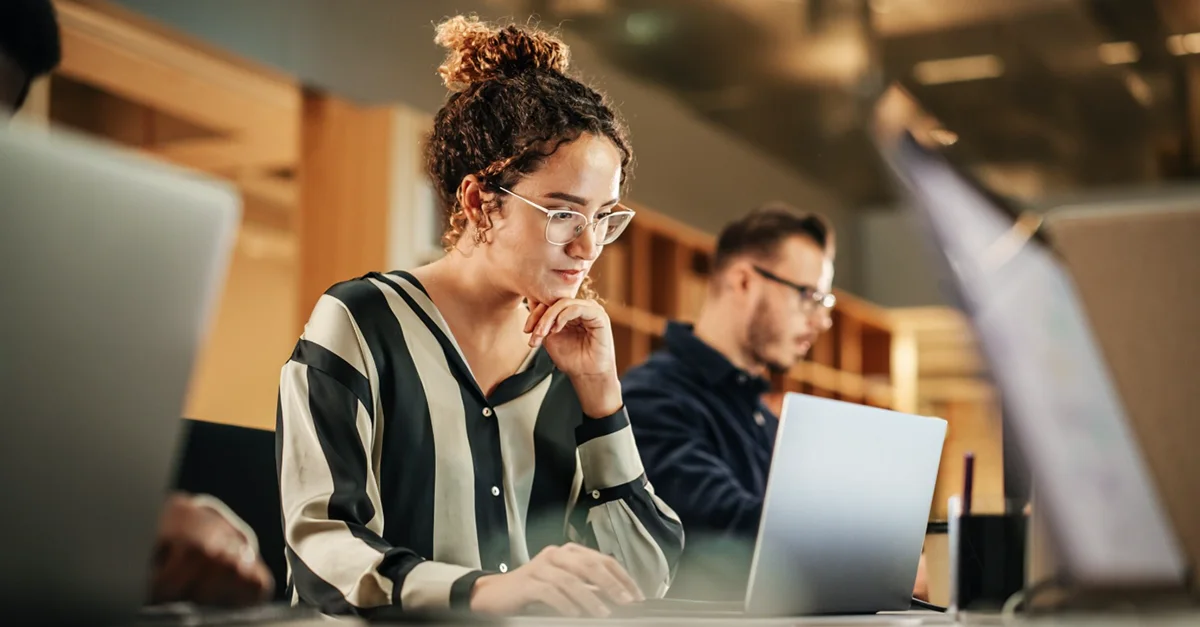 Individual seated at a desk using a laptop in a modern open office, with other people working in the background.