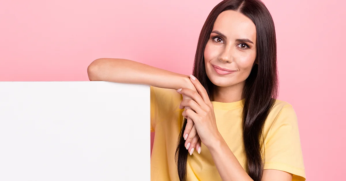 Individual standing beside a large blank white board, resting an arm on top of it while positioned in front of a pink background.
