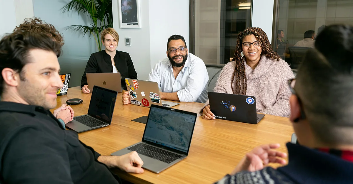 Group of coworkers seated around a table in an office, using laptops and engaging in a collaborative discussion, with multiple devices and stickers visible on the laptop covers.