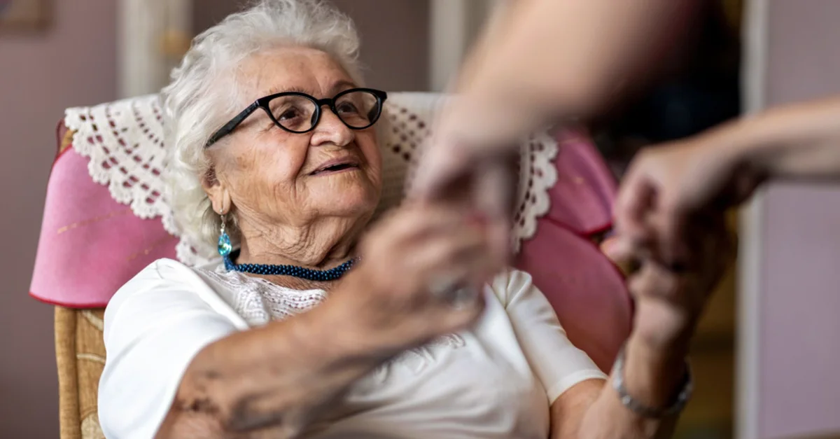 An elderly individual seated in a chair holding hands with another person who is offering support in a home setting.