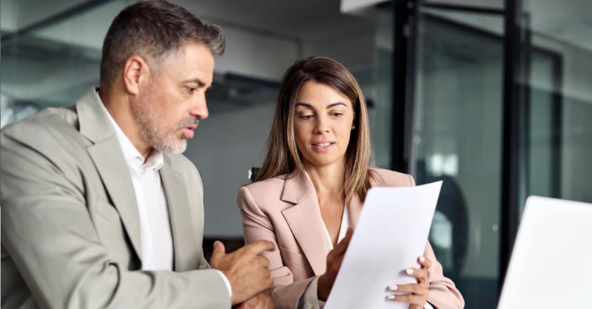 Two colleagues sitting at a desk in an office, reviewing a document together with a laptop open in front of them.