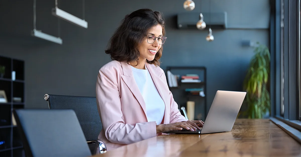 Professional sitting at a desk and working on a laptop in a modern office environment with shelves and workspace decor in the background.