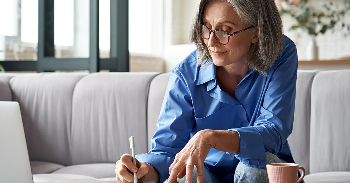 Individual sitting on a sofa with a laptop, holding papers and writing notes with a mug placed nearby.