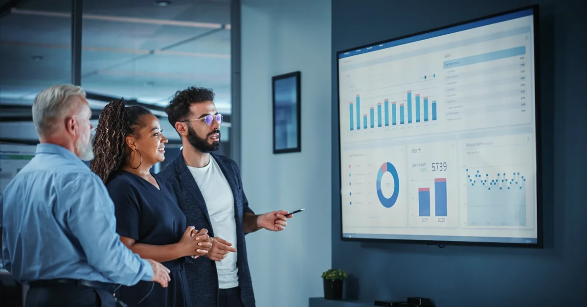 A group of people standing in a modern office, reviewing charts and graphs displayed on a large wall‑mounted screen during a presentation.