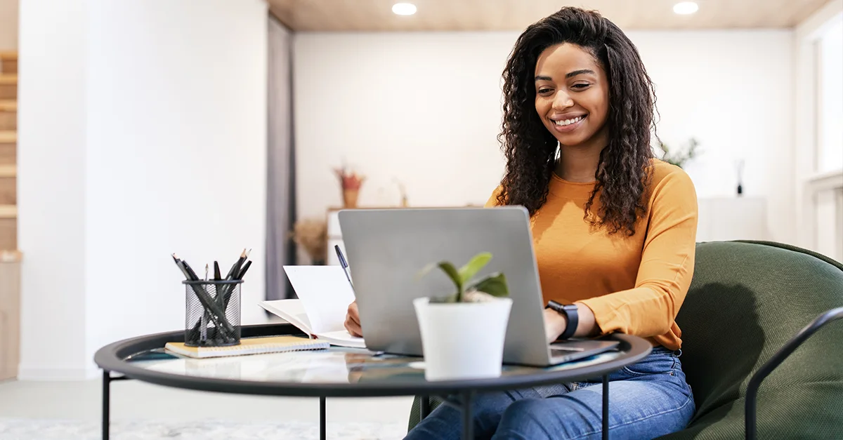 Individual sitting at a round table in a modern home office, using a laptop with documents, stationery, and a small potted plant on the table.