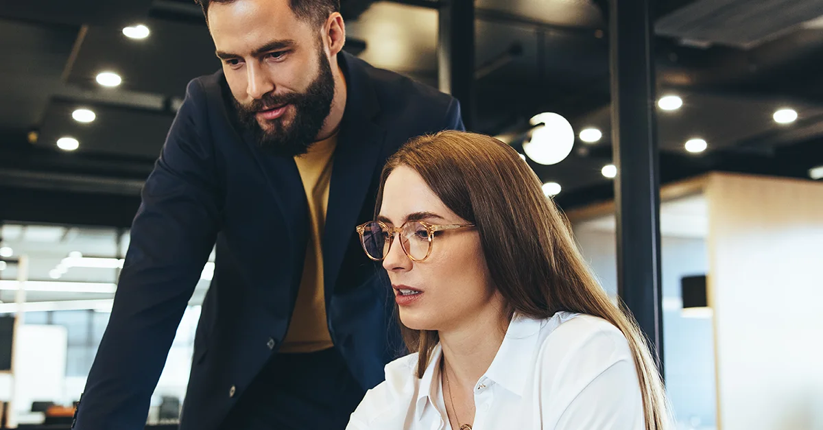 Two coworkers at a desk in a modern office, with one leaning over and pointing while the other looks at a laptop screen, collaborating on a task.