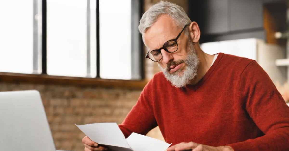 Individual sitting at a desk using a laptop and reviewing paper documents while performing calculations.