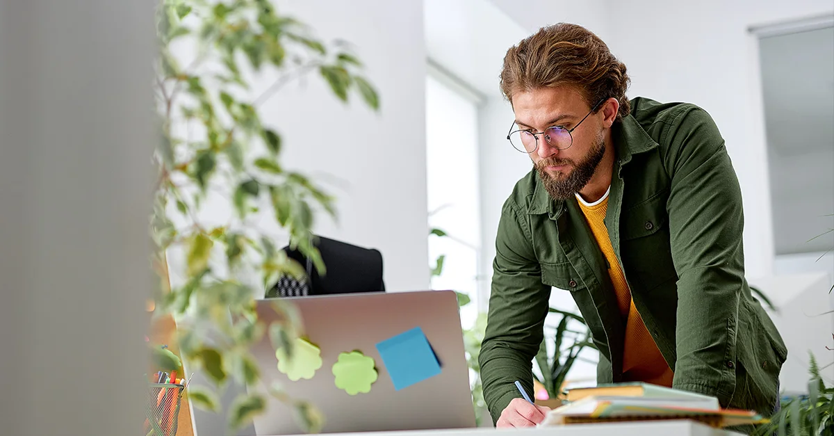Individual standing at a desk, writing in a notebook while working near a laptop with sticky notes on it in a bright office environment.