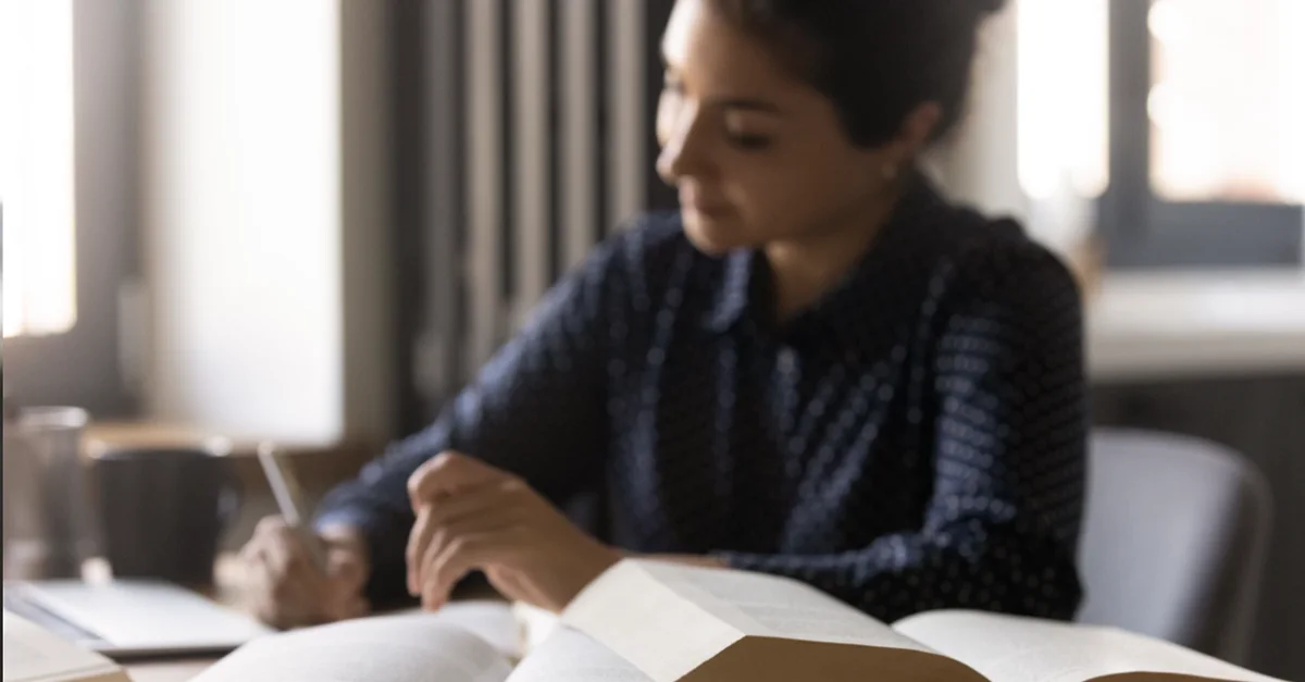 Individual sitting at a desk with several open books, writing notes while studying in a quiet indoor setting.
