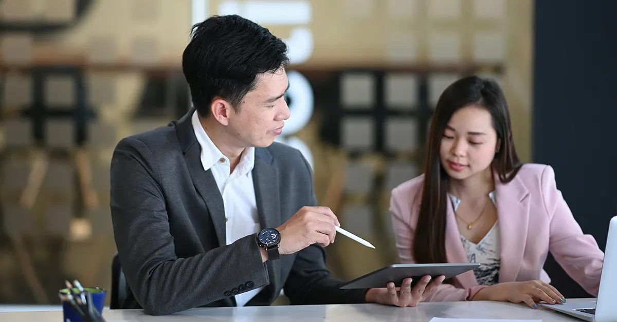 Two professionals sitting at a table during a business meeting, reviewing information on a tablet and taking notes.
