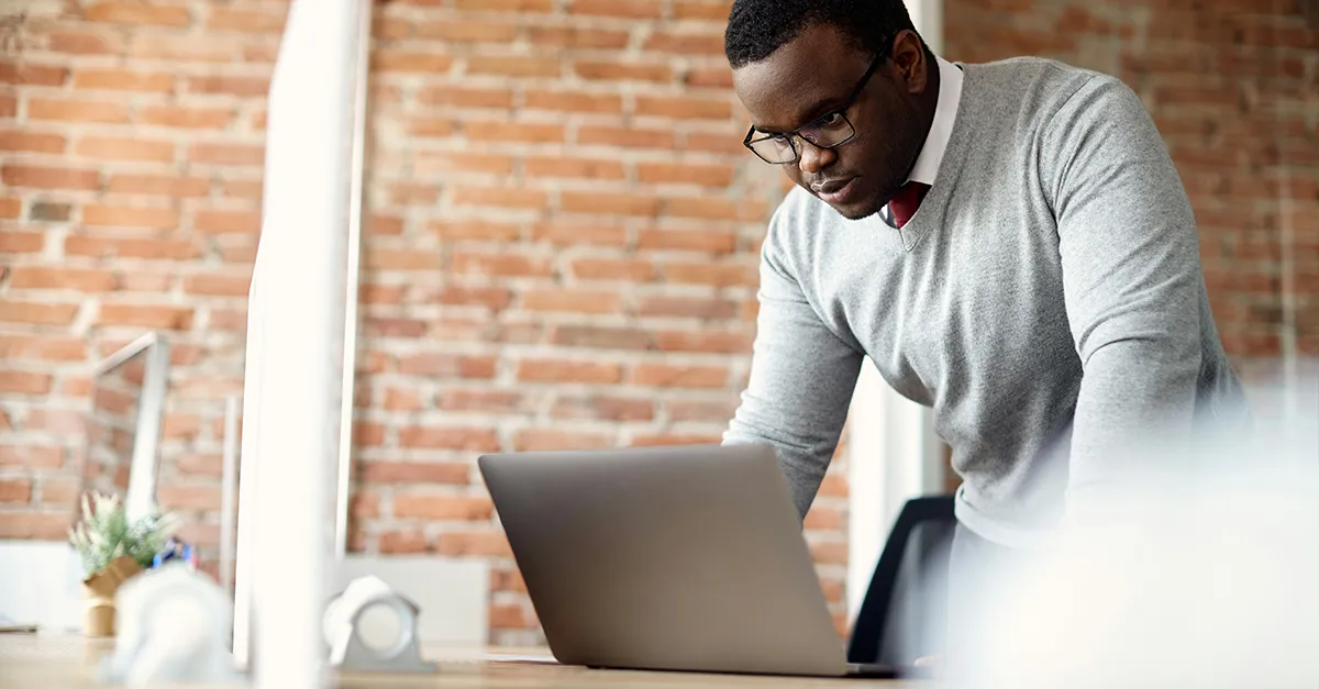 Individual standing at a desk and working on a laptop in a modern office with a brick wall background.