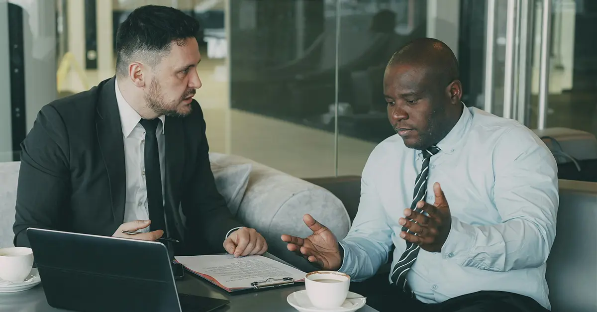 Two professionals sitting at a table in a modern office, discussing documents with a laptop and coffee cups on the table.