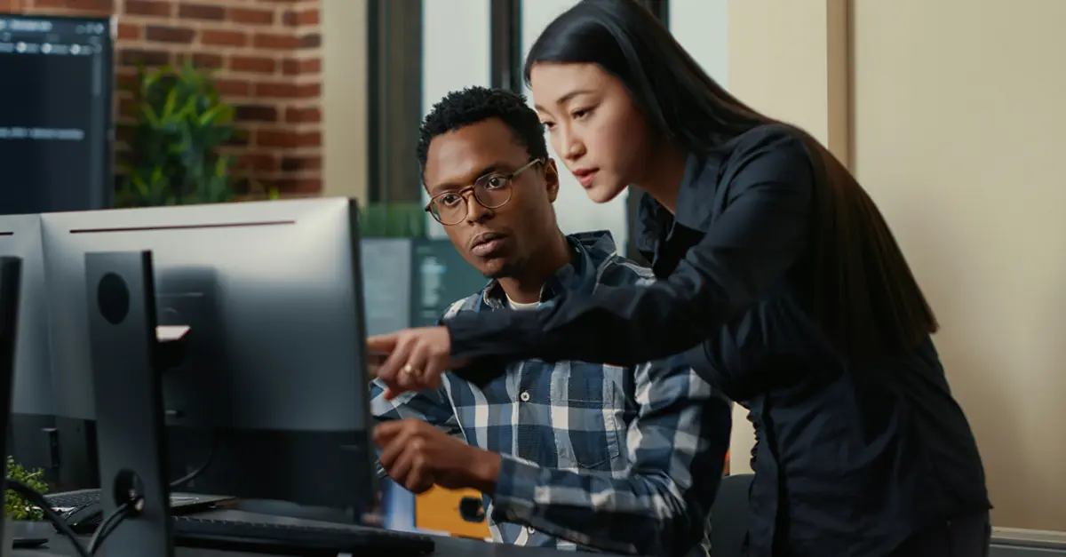 Two colleagues working together at a desk, reviewing information on a computer monitor while discussing and pointing at the screen.