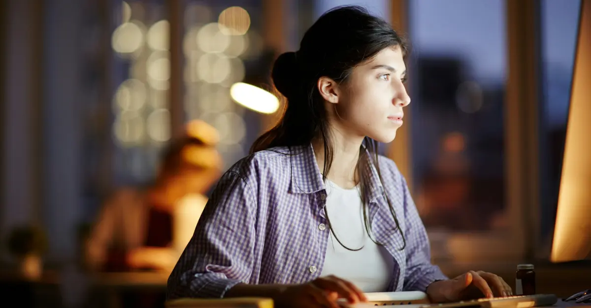 Individual sitting at a desk in a dimly lit office, working on a computer with a lamp in the background.