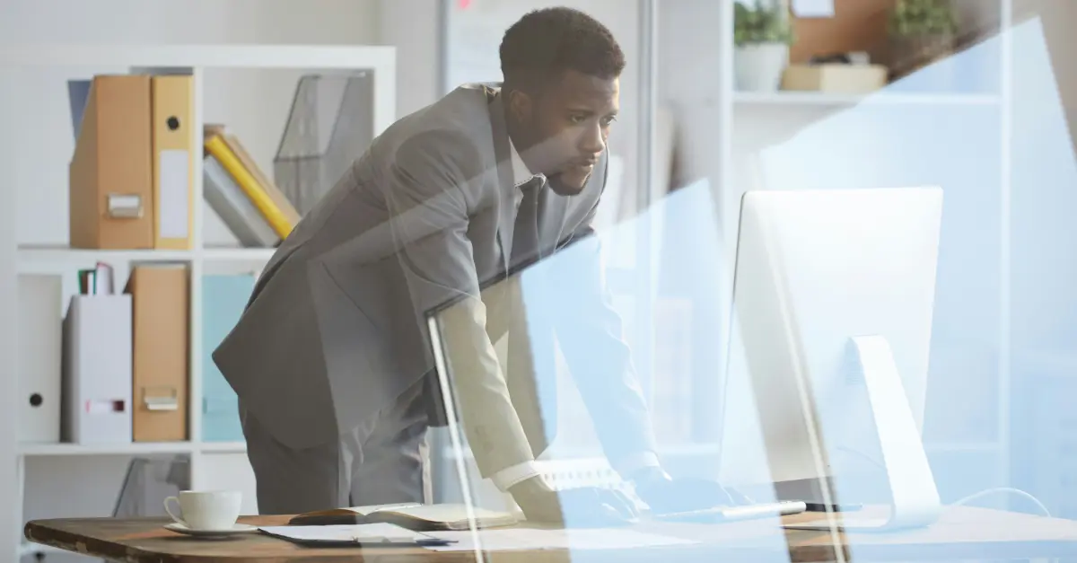 Person in a formal suit standing at an office desk, working on a desktop computer with shelves and folders in the background.