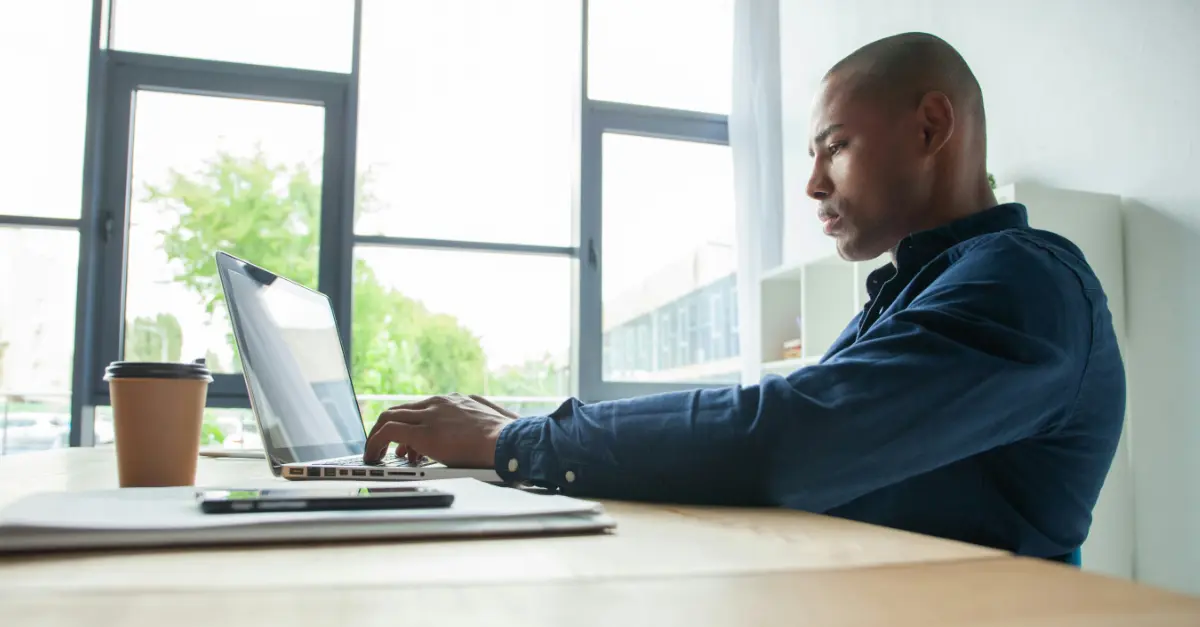 Individual sitting at a wooden desk in a bright office, typing on a laptop with a coffee cup and documents nearby.