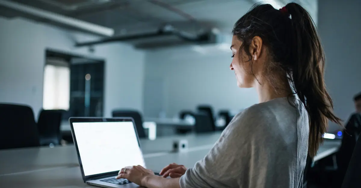 Individual sitting at a desk in a modern office environment, typing on a laptop with multiple chairs and tables in the background.