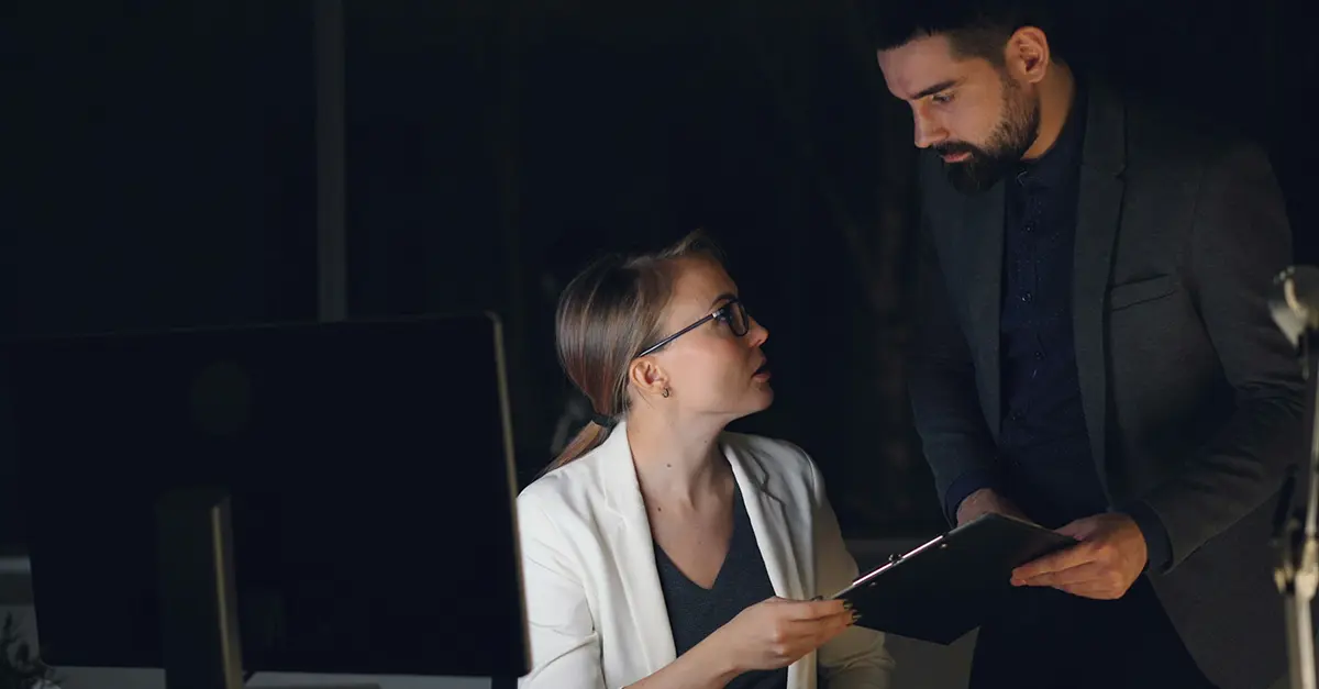 Two colleagues in a dimly lit office, standing and sitting at a desk while reviewing a document with a computer and coffee cup nearby.