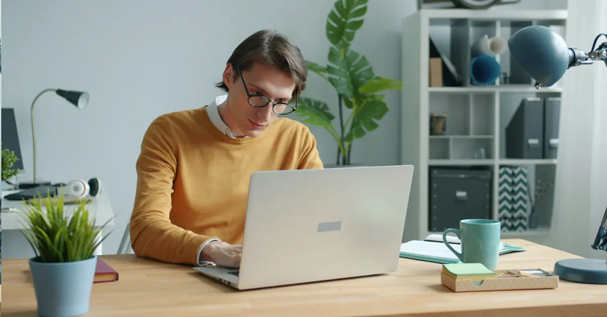 Individual working on a laptop at a wooden desk in a home office with plants, books, and a coffee mug.