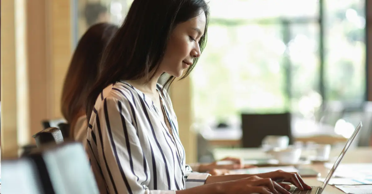 Individual typing on a laptop at a wooden table in a modern cafe with large windows and natural light.