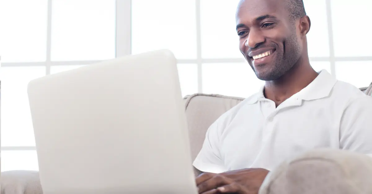 Individual sitting on a beige couch using a white laptop in a bright room with large windows.