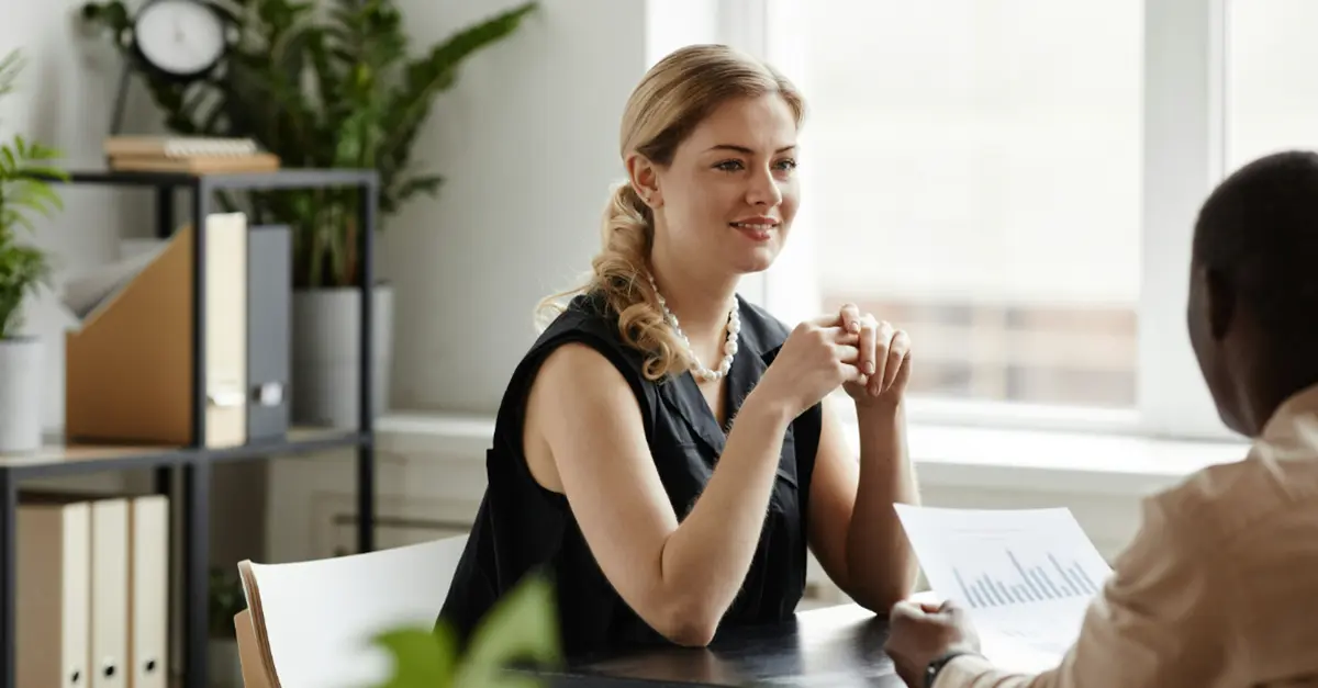 Two individuals seated at a desk in a bright office discussing printed documents with charts, with shelves and plants in the background.