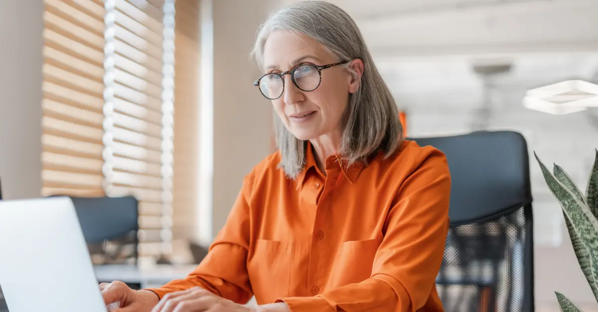 Individual wearing an orange shirt typing on a laptop at a white desk in a bright office with documents and a plant nearby.