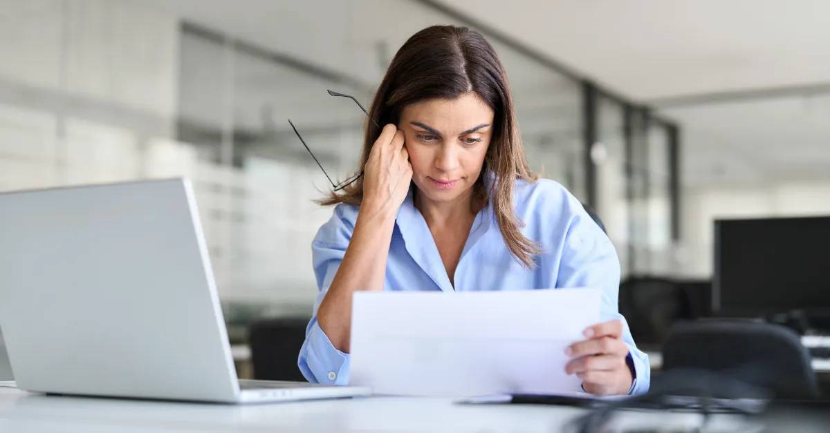 Individual seated at an office desk holding printed documents while working on a laptop, with a modern glass-partitioned background.