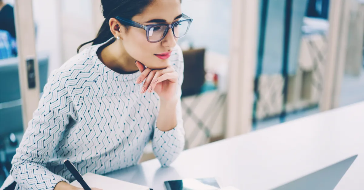 Individual seated at a desk writing in a notebook while working on a laptop, with a smartphone placed nearby in a bright office setting.