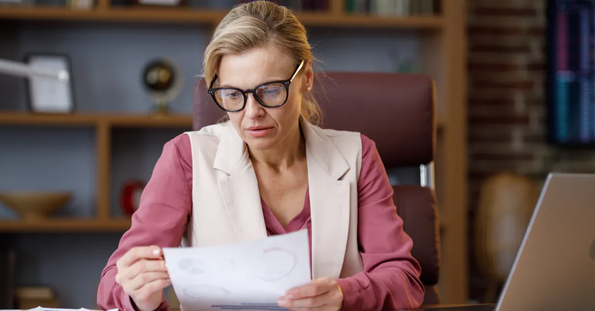 Individual seated at an office desk reviewing printed documents with a laptop and calculator, bookshelves in the background.