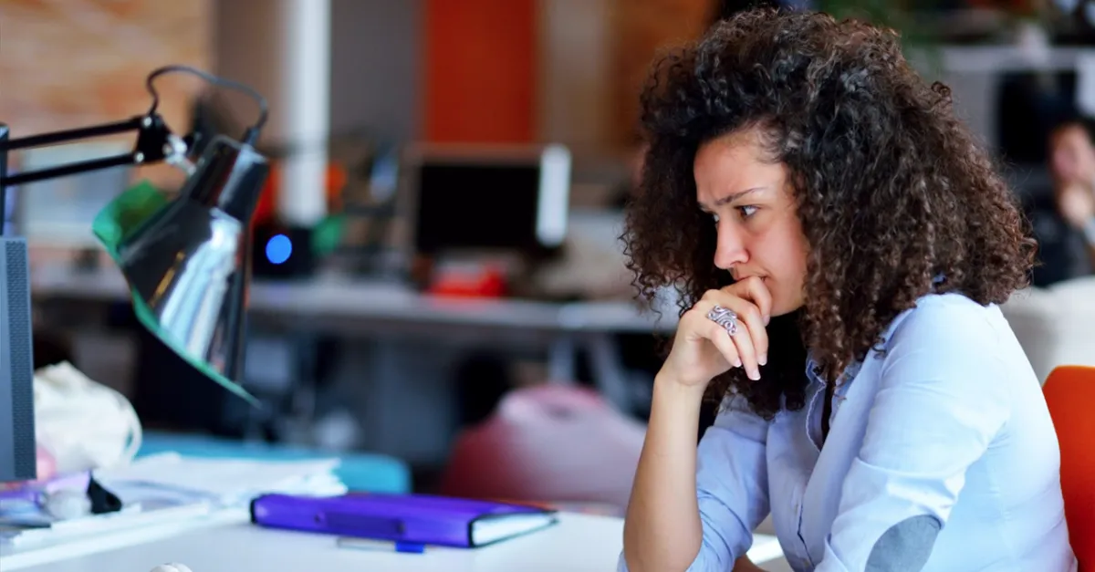 Individual seated at a desk in an open office environment with a notebook, headphones, desk lamp, and office supplies visible.