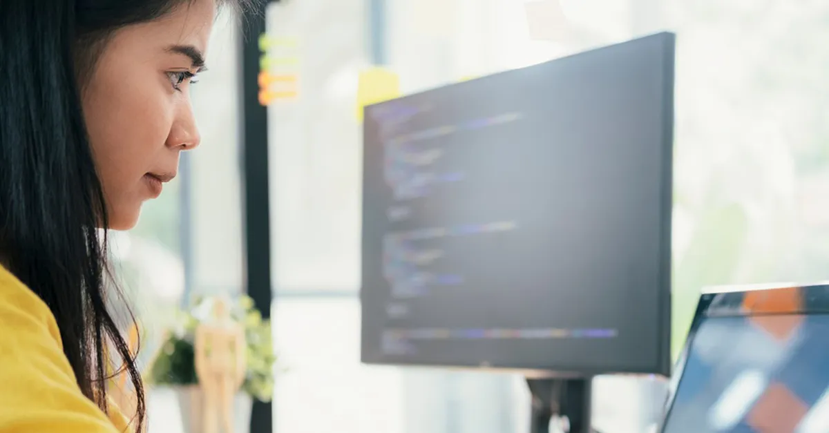 Individual working on a computer with programming code displayed on a monitor in a bright modern office setting.