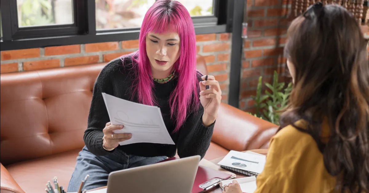 Two individuals seated at a round wooden table in a casual office environment, reviewing printed documents with a laptop and notebooks present.