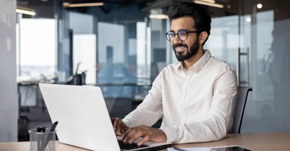 Individual seated at a desk typing on a laptop in a modern office with glass partitions, a notebook, pen, and tablet placed on the table.
