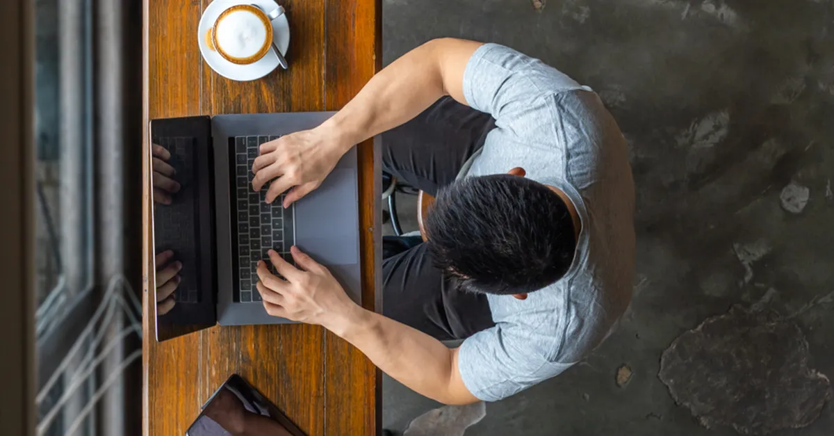 Top view of an individual typing on a laptop at a wooden table with a tablet and a cup of coffee placed nearby.