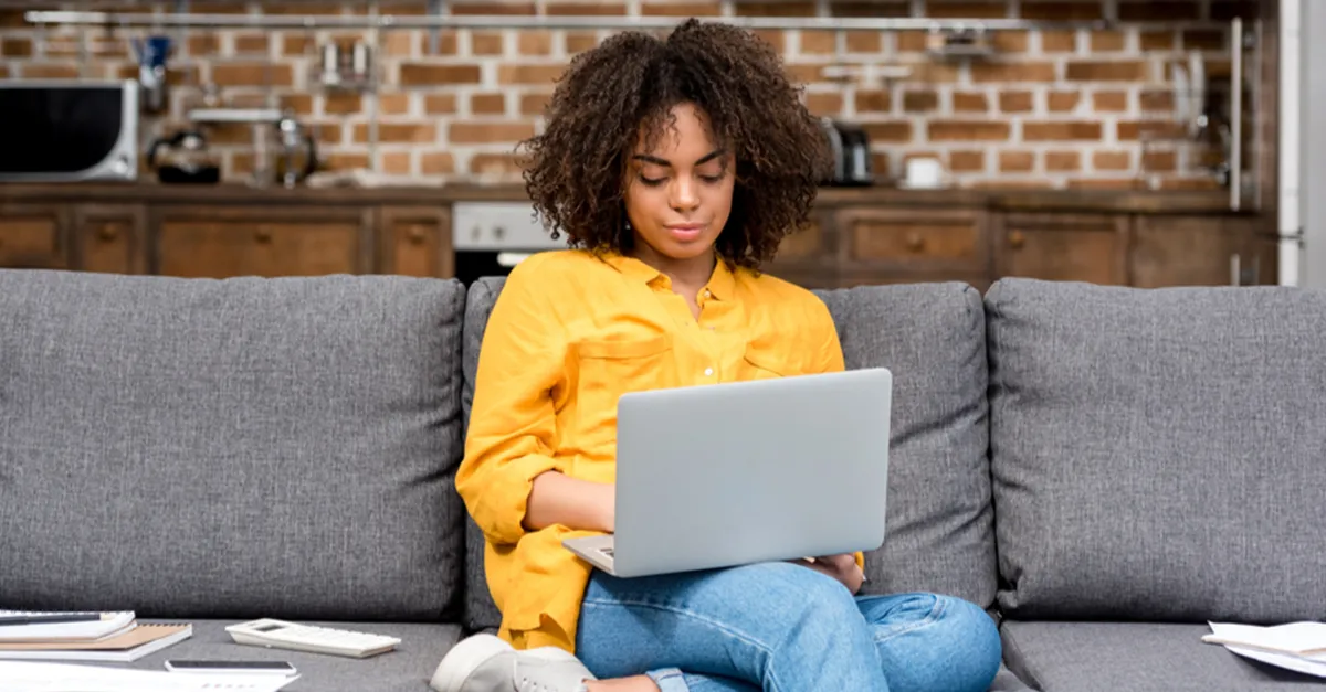 Individual sitting on a gray sofa using a laptop in a cozy home setting with a wooden kitchen background, scattered documents, and a calculator on the table.